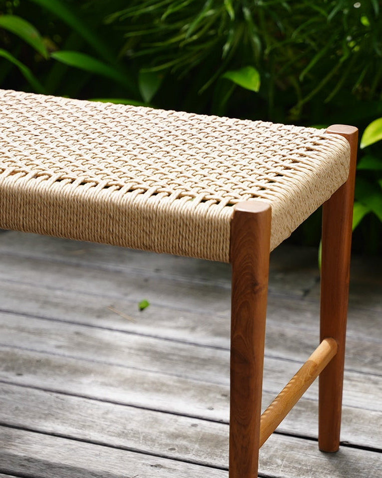 A teak outdoor bench with a woven synthetic seat, placed on a weathered wooden deck with lush green plants in the background.